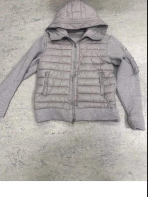 Grå hoodie med quiltad framsida - Grå hoodie med quiltad, vadderad framsida och stickade ärmar samt rygg. Utrustad med dragkedja, två fickor fram och en stor huva. Materialmix av mjuk stickning och polyester, perfekt för en avslappnad och trendig look.