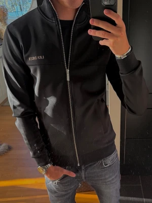 Svart ziptröja från Les Deux - Snygg svart ziptröja från Les Deux med diskret logga på bröstet. Tröjan har hög krage, hel dragkedja framtill och två fickor. Tillverkad i ett mjukt och stretchigt material som ger en sportig look. Perfekt till jeans eller joggers.