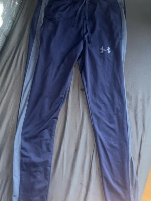 Marinblå träningsbyxor Under Armour - Snygga marinblå träningsbyxor från Under Armour med grå sidopaneler och dragsko i midjan. Tillverkade i mjukt och stretchigt polyestermaterial som passar perfekt för gymmet eller löprundan. Under Armour-logga på vänster lår.