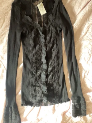Svart ribbad kofta med spetsdetaljer - Supersnygg svart ribbad kofta med knappar framtill och dekorativa spetsdetaljer längs hela framsidan och vid ärmsluten. Djup urringning och långa ärmar ger en feminin och trendig look. Perfekt att styla lager på lager.