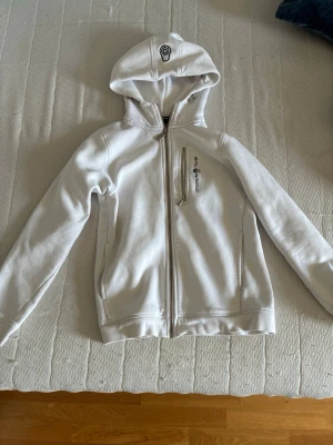 Vit hoodie från Sail Racing - Snygg vit hoodie från Sail Racing med dragkedja framtill och en bröstficka med logga. Hoodien har en broderad detalj på huvan och två sidofickor. Tillverkad i mjukt material som känns skönt mot huden. Perfekt för en avslappnad och sportig stil.