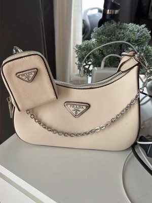 Beige axelremsväska från Prada - Snygg beige axelremsväska från Prada med matchande liten väska. Väskan har silverfärgade detaljer, kedja och dragkedja samt det klassiska Prada-emblemet framtill. Tillverkad i canvasliknande material och har en modern, stilren form.