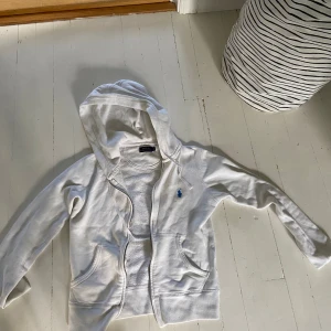 Vit hoodie från Polo Ralph Lauren XS - Vit hoodie från Polo Ralph Lauren i mjuk bomull. Klassisk design med dragkedja, huva och två fickor framtill. Liten blå broderad logga på bröstet. Perfekt för chill dagar eller när du vill ha en clean och enkel look. Tveka inte att fråga!