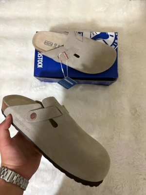 Taupe Birkenstock Boston storlek 42 - Snygga taupe tofflor från Birkenstock i mjuk mocka med klassisk design och justerbart spänne i metall. Yttersulan är i mörkbrunt gummi med vågigt mönster för bra grepp. Perfekta för chill dagar hemma eller när du vill ha det bekvämt.