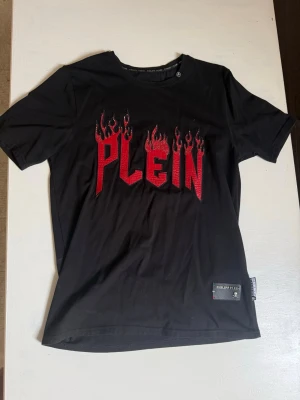 Svart Philipp Plein t-shirt med flames - Svart t-shirt från Philipp Plein med coolt rött flames-tryck och texten 'PLEIN' på bröstet. Klassisk passform och rund hals. Liten logga på nedre kanten och diskret emblem i nacken. 