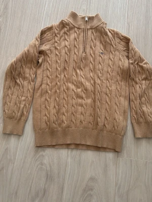 Gant kabelstickad tröja  - Säljer en beige kabelstickad tröja med halv dragkedja och hög krage. Tröjan har långa ärmar, ribbade muddar och nederkant samt ett diskret broderat märke på bröstet. Perfekt för dig som gillar klassisk och stilren look.