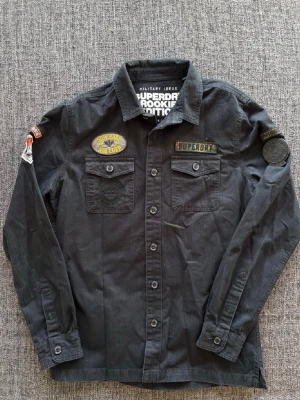 Svart overshirt med patches från Superdry - Säljer en svart långärmad overshirt från Superdry Rookie Edition med flera coola patches och bröstfickor med knappar. Overshirten har militärinspirerad stil och är gjord i bomull. Perfekt för dig som gillar streetwear och detaljer. Storlek L, passar M