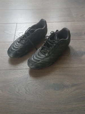 Svarta klassiska fotbollsskor - Säljer ett par svarta fotbollsskor med snörning och låg profil. Skorna har en stilren design med diskreta sömmar och dobbar för bra grepp på planen. Perfekta för dig som vill ha en enkel och funktionell sko till match eller träning.låda ingår.