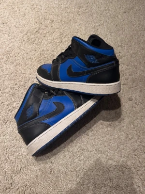Nike Air Jordan 1 Mid svart/blå - Säljer ett par Nike Air Jordan 1 Mid sneakers i svart och blått läder med vita sulor och klassisk Swoosh på sidan. Skorna har ikoniska Air Jordan-detaljer och en stilren design som passar till det mesta. Perfekta för dig som gillar streetwear och vill sticka ut.