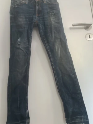 Mörkblå slitna Nudie jeans - Säljer ett par mörkblå Nudie jeans med slitningar. Jeansen har klassisk femficksmodell och kontrastsömmar. Jeansens längd sitter mer som 28-30 än 32