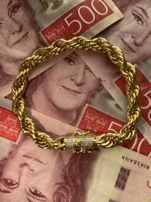 Guldpläterat Cordell Armband med Diamantlås  - Säljer ett chunky armband i guldfärgad metall med tvinnad repdesign. Låset är dekorerat med små gnistrande stenar som ger extra bling. Perfekt för dig som gillar statement-smycken och vill sticka ut lite extra.
