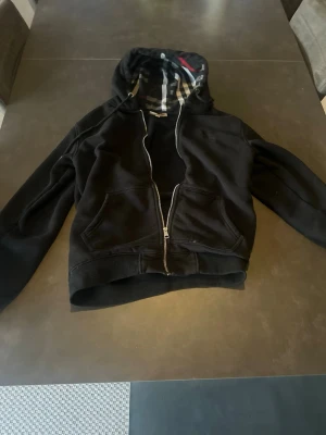 Svart hoodie med dragkedja och huva - Svart hoodie med dragkedja framtill och två fickor. Huvan har ett rutigt innerfoder i svart, vitt och rött som ger en snygg kontrast. Klassisk passform och ribbade muddar vid ärmar och nederkant. Perfekt för en avslappnad och stilren look.