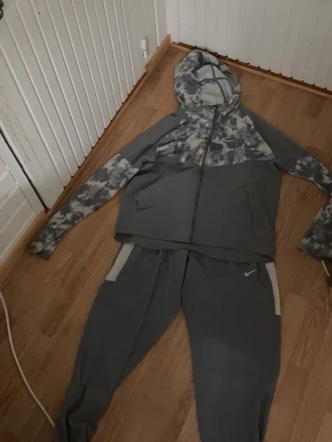 Grå Nike träningsset med camomönster - Säljer ett grått träningsset från Nike med zip-hoodie och matchande byxor. Hoodien har camomönster över axlar och huva samt fickor framtill. Byxorna har dragkedjor vid benslut och vita detaljer vid fickorna. Perfekt för träning eller chill.