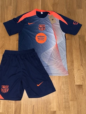 FC Barcelona träningsset  - Säljer ett snyggt FC Barcelona träningsset med t-shirt och shorts i marinblått med orange detaljer. Helt ny