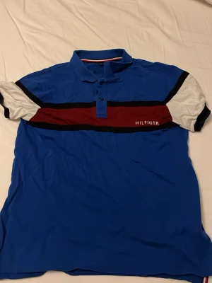Blå piké från Tommy Hilfiger - Snygg blå pikétröja från Tommy Hilfiger med vita och röda detaljer samt svarta ränder över bröstet. Klassisk krage och knappar framtill. Perfekt för en avslappnad men stilren look.