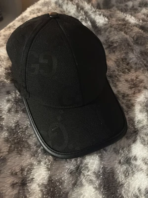 Svart keps med GG-mönster - Snygg svart keps med diskret GG-mönster över hela ytan. Kepsen har böjd skärm med läderliknande kant och är tillverkad i ett textilmaterial. Perfekt accessoar för en trendig streetstyle-look.