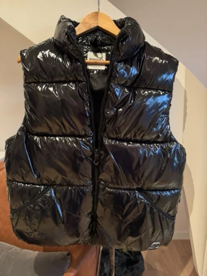Svart glansig dunväst från Bershka - Snygg svart dunväst från Bershka med glansig finish och hög krage. Västen har dragkedja framtill och två fickor på sidorna. Perfekt för lager på lager och ger en cool streetwear-look. Materialet är syntet med dunfyllning.