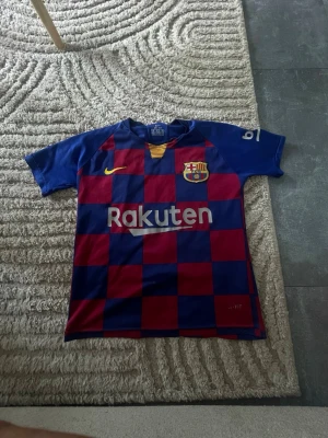Barcelona matchtröja Nike blå/röd - Säljer en Barcelona matchtröja från Nike i blått och rött rutmönster. Tröjan har klubbmärke, Rakuten-tryck på bröstet och gula detaljer vid halsen. Tillverkad i Dri-FIT material som andas, perfekt för fotboll eller träning.