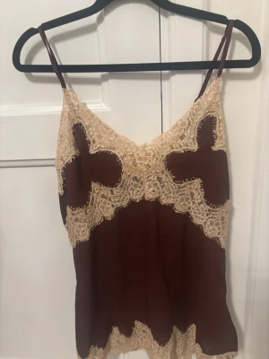 Viral Zara linne - Snyggt brunt linne med tunna axelband och dekorativa beige spetsdetaljer längs urringning, nederkant och mittparti. Perfekt för en chic och avslappnad look, med en feminin touch tack vare spetsen. Materialet ser ut att vara mjukt och följsamt.