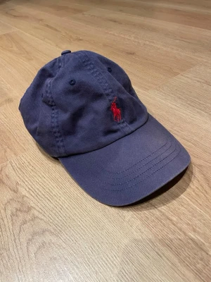 Mörkblå keps från Polo Ralph Lauren - Snygg mörkblå keps från Polo Ralph Lauren med klassisk röd broderad logga framtill och 'POLO' broderat bak. Justerbart spänne i metall baktill för perfekt passform. Tillverkad i bomullstyg som andas och passar till de flesta outfits.