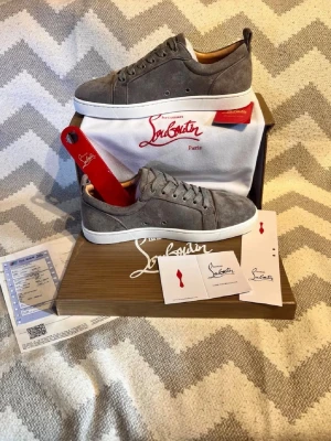Gråa Louboutin sneakers i mocka - Snygga gråa sneakers från Christian Louboutin i mjuk mocka med klassisk vit sula och ikonisk röd undersida. Skorna har lågt skaft, rund tå och matchande grå skosnören. Perfekta för dig som vill sticka ut med exklusiv stil.