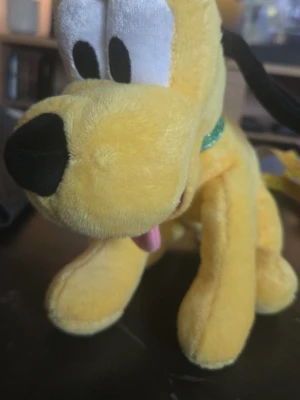 Gul Pluto mjukisdjur - Säljer ett gulligt mjukisdjur som föreställer Pluto från Disney. Gosedjuret är gult med svart nos, svarta öron och en grön glittrig halsband. Tungan sticker ut och ögonen är stora och vita med svarta pupiller. Perfekt som present eller dekoration!