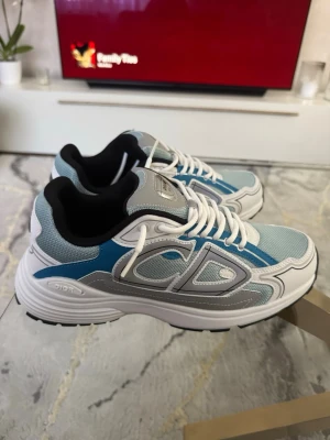 Dior B30 sneakers blå och vit - Säljer ett par Dior B30 sneakers i blått, vitt och grått med stora CD-detaljer på sidan. Skorna har mesh och läderpaneler, vita snören och svart sula. Snygg och sportig design som sticker ut och passar perfekt till streetwear.