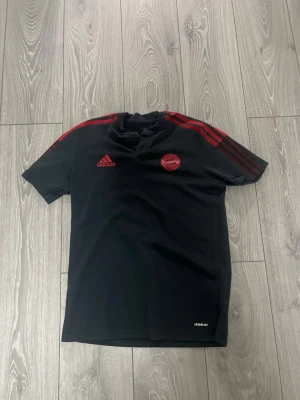 Svart Bayern München t-shirt Adidas - Snygg svart t-shirt från Adidas med Bayern München-logga på bröstet och röda detaljer på axlarna. Tillverkad i Primegreen-material och har knappslå vid halsen. Perfekt för dig som gillar sportig stil och fotboll.