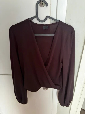 Vinröd omlottblus från Gina Tricot - Snygg vinröd omlottblus från Gina Tricot med långärmad design och djup v-ringning. Blusen har ett mjukt, lätt material och resår vid ärmsluten för en trendig look. Perfekt att styla med jeans eller kjol.