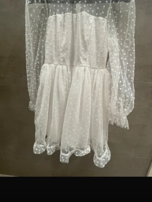 Vit kort klänning med hjärtan - Endast använd vid ett tillfälle! En Supersöt vit kort klänning med transparenta långärmar och små hjärtan som mönster. Klänningen har puffiga ärmar med resår vid handlederna och volangkant nedtill. Tillverkad i ett lätt, syntetiskt material som ger en romantisk look.