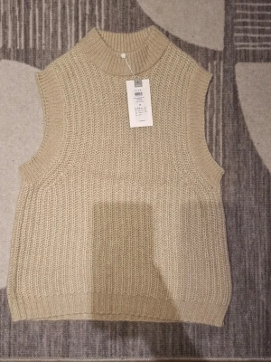 Beige stickad väst från Vila - Säljer en beige stickad väst från Vila med hög krage och grov ribbad struktur. Västen är ärmlös och har en avslappnad passform, perfekt att styla över en skjorta eller t-shirt. Enkel och stilren design som passar till många outfits.