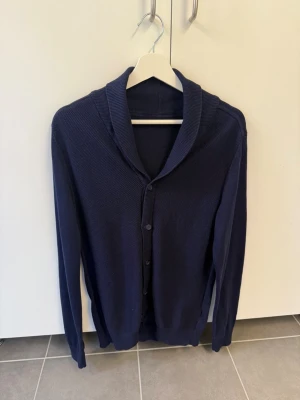 Mörkblå kofta från Selected Homme, stl M - Stilren mörkblå kofta från Selected Homme med sjalkrage och knäppning framtill. Tillverkad i mjukt, tunt stickat material som passar perfekt till lager på lager. Långärmad modell med enkel design som är lätt att matcha.