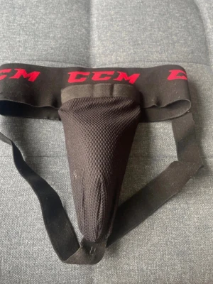 Svart suspensoar från CCM - Svart suspensoar från CCM med bred resår i midjan och röda logotyper. Skyddsdelen är tillverkad i ventilerande meshmaterial för extra komfort vid sportaktiviteter. Perfekt för hockey eller andra kontaktsporter.