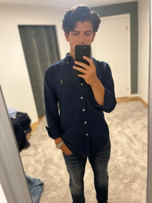Mörkblå skjorta från Ralph Lauren - Klassisk mörkblå skjorta från Ralph Lauren med button-down krage och broderad logga på bröstet. Skjortan har långa ärmar, vita knappar och är tillverkad i bomull. Perfekt för en stilren och avslappnad look. Tveka inte på att skriva om du har frågor eller funderingar🌟😊