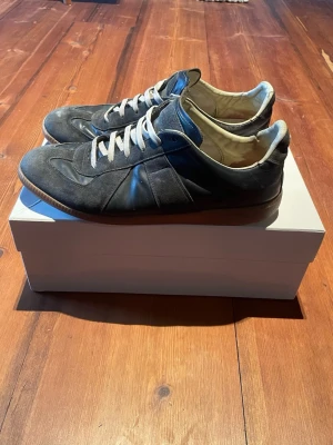 Maison margiela gats - Säljer ett par svarta sneakers med vita skosnören och bruna sulor. Skorna är låga och har en snygg mix av mocka och läder på ovandelen. Insidan är ljus och de har en klassisk, stilren look som passar till det mesta.