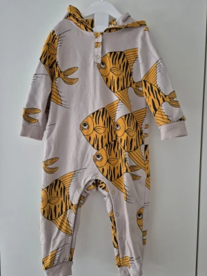 Mini rodini onesie 80-86 - En långärmad onesie från Mini Rodini med huva och tryckknappar framtill och vid benen. Plagget har ett stort, färgglatt fiskmönster i gult och svart. Muddar vid ärm- och benslut för extra komfort. Perfekt för dig som gillar lekfulla prints. Sparsamt använd trots sina år på nacken. Fortfarande härlig i tyget. Ett riktigt guldkorn.