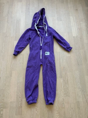 Lila onepiece med huva och dragkedja - Mysig lila onepiece med huva och vita dragsnören. Dragkedja framtill och muddar vid ärmar och benslut för extra komfort. Perfekt för slappa dagar hemma. Tillverkad i mjukt sweatshirtmaterial. Liten patchdetalj på ena sidan.