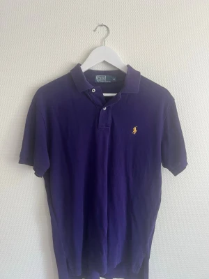 Lila pikétröja från Polo Ralph Lauren - Säljer en klassisk lila pikétröja från Polo Ralph Lauren med gul broderad logga på bröstet. Tröjan har korta ärmar, krage och knappar vid halsen. Perfekt för en avslappnad men ändå stilren look.