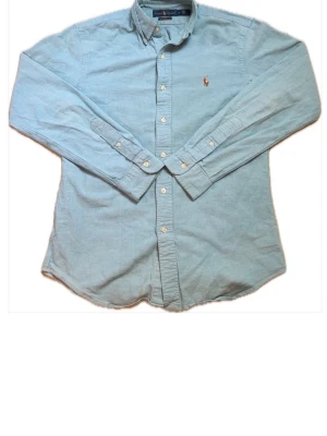 Ljusblå skjorta från Ralph Lauren - Klassisk ljusblå skjorta från Ralph Lauren i slim fit-modell. Tillverkad i bomull med stretch för extra komfort. Skjortan har knappar framtill, button-down-krage och den ikoniska broderade loggan på bröstet. Perfekt till jeans eller chinos.