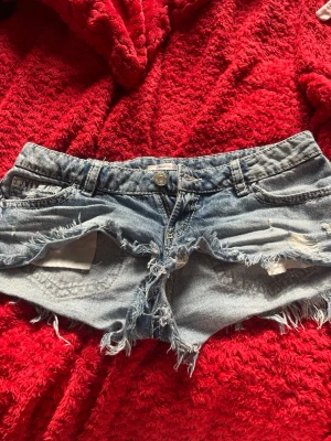 Ljusblå  jeansshorts - Säljer ett par ljusblå jeansshorts med råa, slitna kanter och fransar. Shortsen har klassisk femficksmodell, knapp och dragkedja framtill samt tydliga slitningar för en edgy look. Perfekta för sommardagar och festivaler.