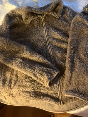 Beige teddyjacka med dragkedja - Mysig beige teddyjacka med hög krage och hel dragkedja framtill. Jackan har en fluffig, lurvig yta och mjuka muddar vid ärmar och nederkant. Perfekt för dig som vill ha en varm och trendig look under kyliga dagar.
