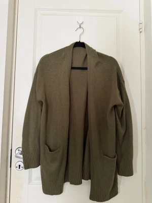 Olivgrön oversized stickad kofta - Mysig olivgrön stickad kofta med två stora fickor framtill och ribbade muddar vid ärmslut och nederkant. Koftan har en avslappnad, oversized passform och är öppen framtill utan knappar. Perfekt för lager på lager och chill stil.