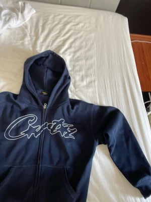 Mörkblå Corteiz hoodie - Snygg mörkblå hoodie med dragkedja och stor huva. Hoodien har fickor på magen och är tillverkad i mjukt material, perfekt för en avslappnad stil.