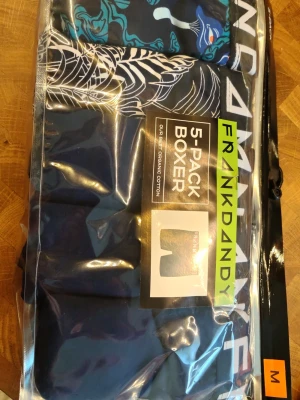 Frank Dandy 5-pack boxerkalsonger M - 5-pack boxerkalsonger från Frank Dandy i storlek M. Flera olika mönster och färger i blåa och svarta toner, vissa med grafiska tryck och logga. Tillverkade i mjuk ekologisk bomull för maximal komfort. Oöppnad förpackning.