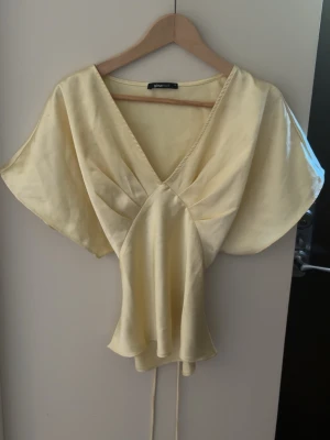 Ljusgul omlottblus i satin - Supersnygg ljusgul omlottblus i glansigt satinmaterial med djup v-ringning och knytband i midjan. Blusen har korta, vida ärmar och ett flowy fall som ger en elegant siluett. Perfekt att styla med jeans eller kjol för en trendig look.