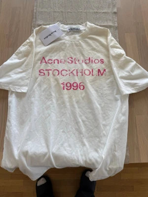Vit Acne Studios t-shirt - Vit t-shirt från Acne Studios med rosa texttryck 'Acne Studios STOCKHOLM 1996' på bröstet. Klassisk passform och rund halsringning. Perfekt för en trendig och avslappnad stil.