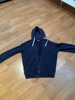 Mörkblå hoodie med dragkedja - Mörkblå hoodie med dragkedja och vita snören i huvan. Klassisk design med två fickor framtill och ribbade muddar vid ärmar och nederkant. Liten röd logga broderad på bröstet. Perfekt för en avslappnad stil.
