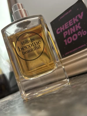 Things Become Beautiful Eau de Parfum - En stilren parfymflaska i genomskinligt glas med silverfärgad spraykork. Flaskan rymmer 50 ml och innehåller en gyllene vätska. Texten 'things become beautiful' står tryckt på framsidan. Eau de Parfum med 80% volymalkohol.