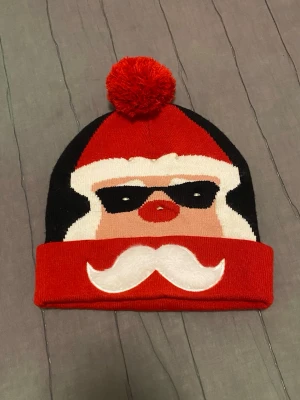 Röd och svart mössa med tomtemotiv - Säljer en stickad mössa i rött och svart med en stor röd tofs på toppen. Framsidan har ett coolt tomtemotiv med solglasögon och vit mustasch. Mössan har uppvikt kant och är perfekt för vintern och julen. Materialet känns mjukt och stretchigt.