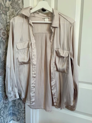 Beige silkeslen skjorta med fickor - Säljer en beige skjorta i silkesliknande material med två bröstfickor och klassisk krage. Skjortan har långa ärmar med knäppning vid manschetterna och en avslappnad passform. Perfekt för dig som gillar stilrena och mjuka plagg.
